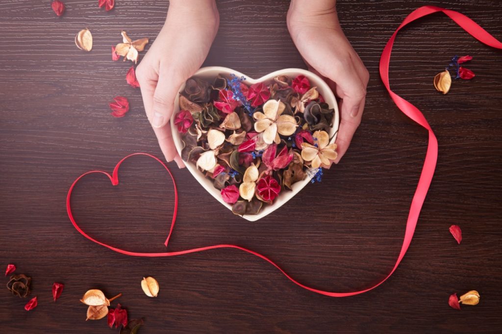 Hands holding a heart shaped bowl of flowers on a wooden table.