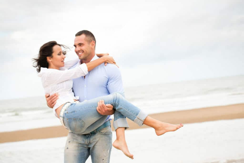 A man is carrying a woman on the beach.