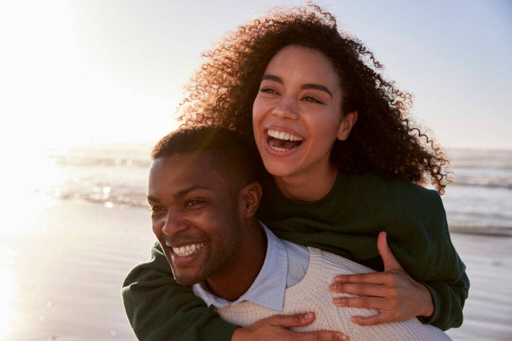 A young black man is carrying a woman on his back at the beach.