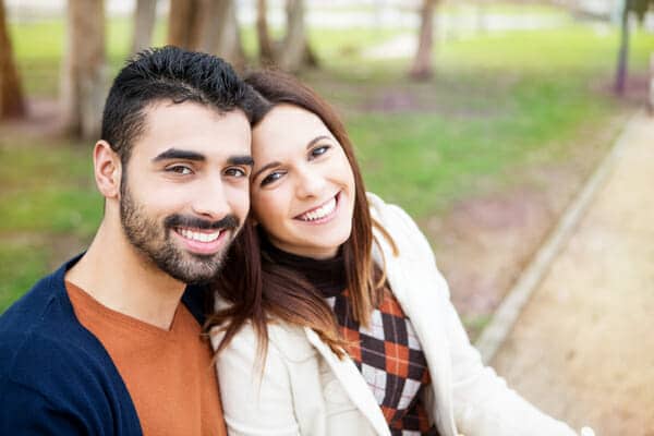 A man and woman are smiling while sitting on a bench in a park.