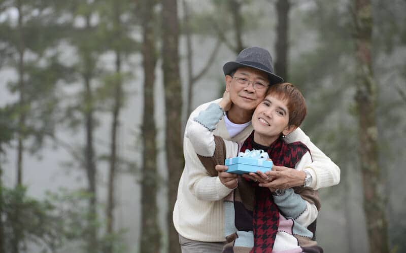 An elderly couple holding a birthday cake in the forest.