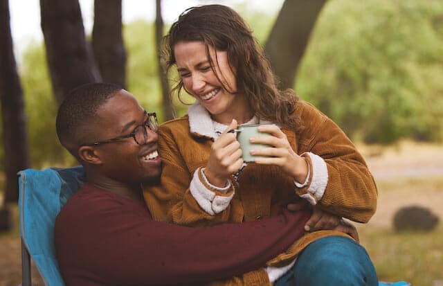 A man and woman are sitting in a chair in a park holding a cup of coffee.