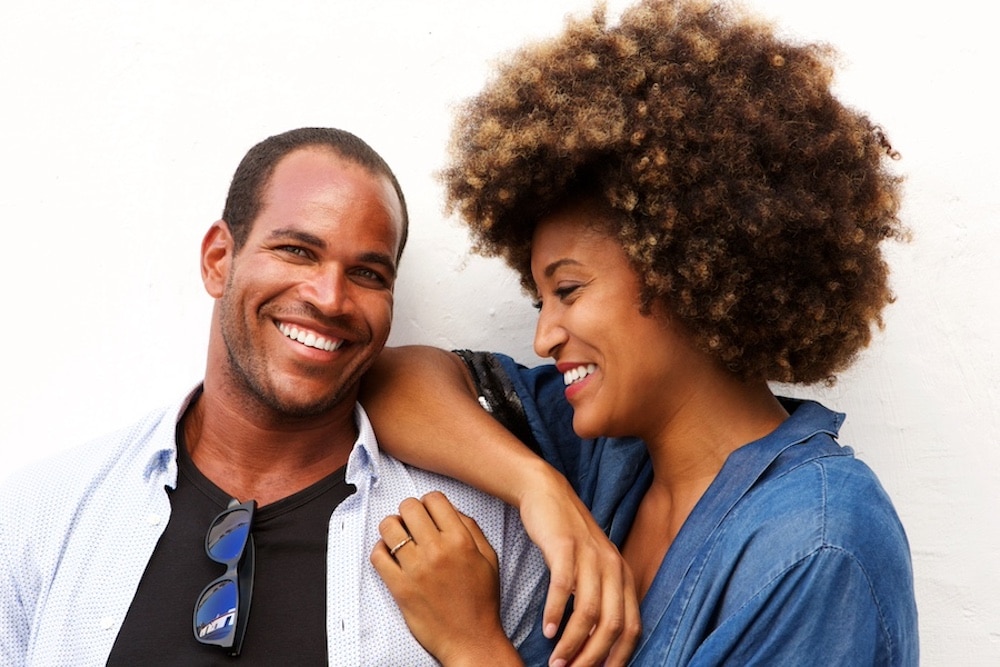 A man and a woman are smiling and standing close together. The woman has her arm on the man's shoulder. Both are casually dressed, and the background is plain white.