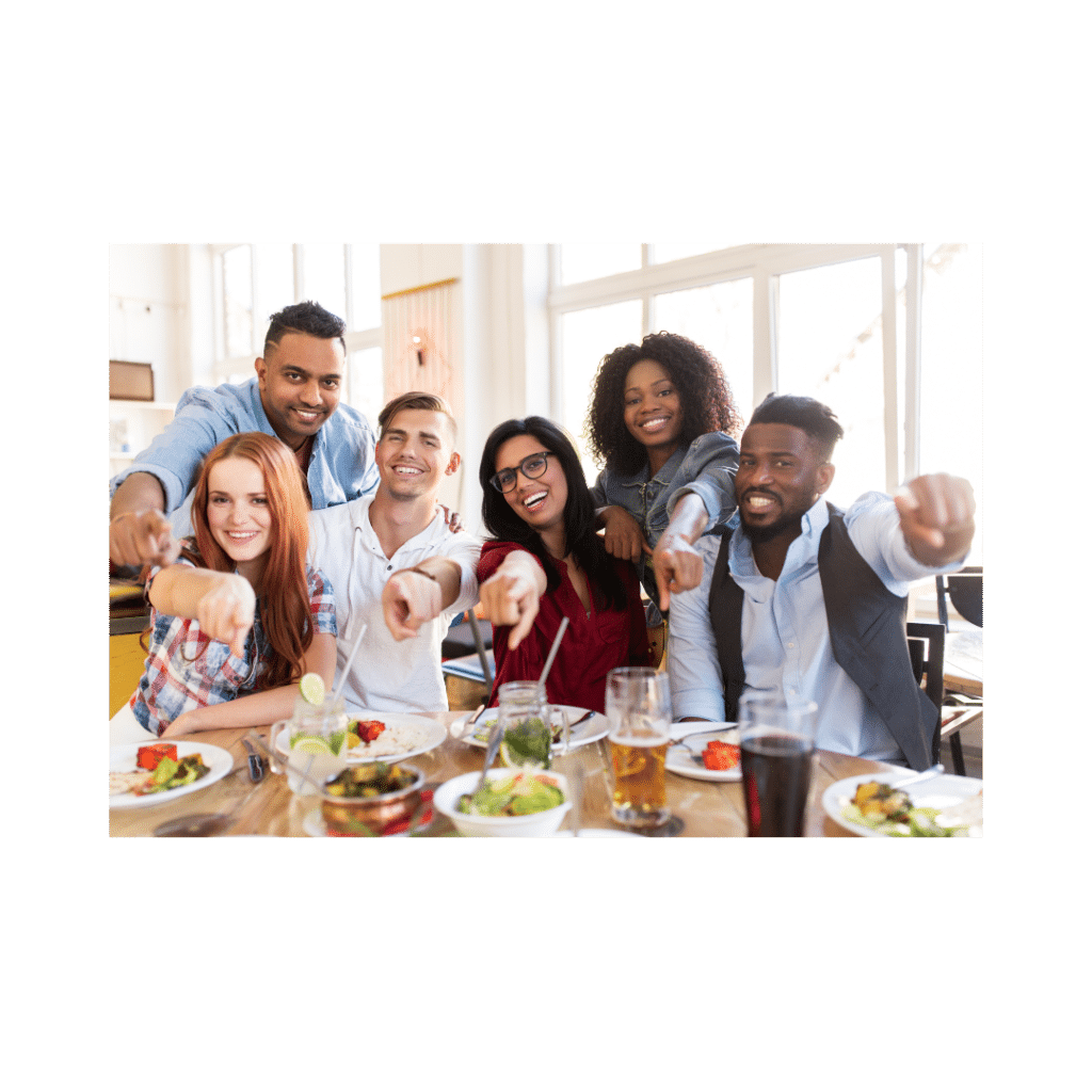 A group of six people sit at a restaurant table enjoying a meal, smiling and pointing towards the camera. Plates of food and drinks are on the table.