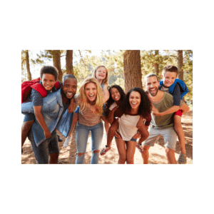 A diverse group of people, including adults and children, smiling and posing together outdoors in a forest setting. Some have children on their backs.