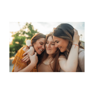 Three women stand close together outdoors with eyes closed and joyful expressions, sharing a warm embrace. Trees and soft sunlight are visible in the background.